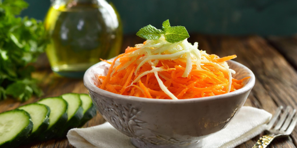 Une salade de carottes et concombre râpés dans un bol. Des tranches de concombre à gauche du bol sur une table en bois et une fourchette à droite. Dans le fond une bouteille en verre d'huile d'olive et du persil sur la gauche. le bol est posé sur une serviette en tissu blanc.